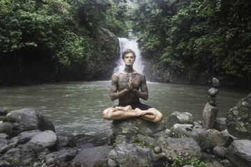 Fototapeta na wymiar tattooed man sitting in lotus position on rock with Aling-Aling waterfall and green plants on background, Bali, Indonesia