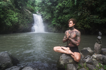tattooed man sitting in lotus position on rock with Aling-Aling waterfall and green plants on background, Bali, Indonesia