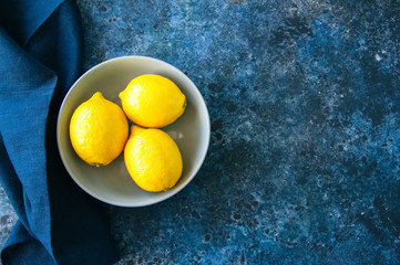 Three ripe yellow lemons in a grey bowl on a blue stone background. Top view and copy space.