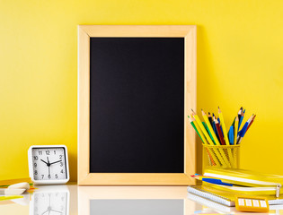 Chalkboard and school supplies on white table by the yellow wall. Side view, empty space for text. Back to school concept.