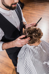 high angle view of curly kid covered with striped cloth sitting at barbershop while barber cutting his hair