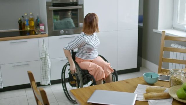 Medium shot of independent paraplegic woman in wheelchair putting away milk bottle into fridge after finishing breakfast