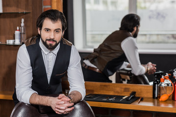 handsome barber leaning on chair at workplace and looking at camera