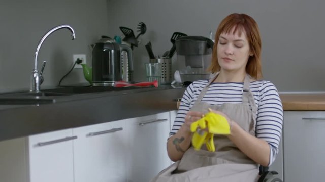 Tracking of cheerful independent woman in wheelchair putting on gloves before washing dishes after breakfast