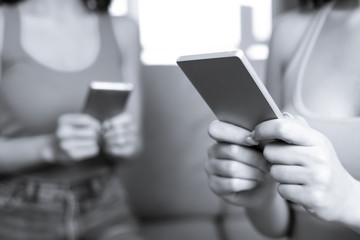 Technology and communication. Two young girl friends sitting on a sofa at home writing messages on the phone or using the Internet.