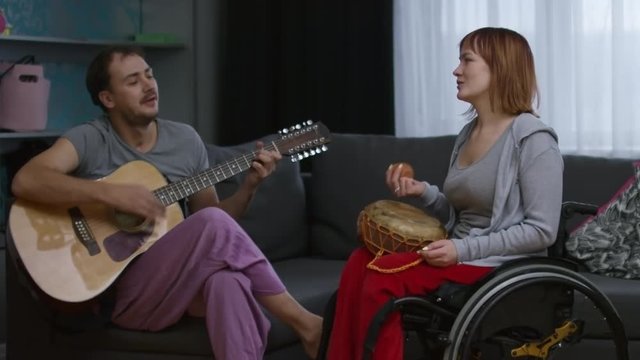 Medium shot of paraplegic happy woman in wheelchair snapping fingers and moving shaker instrument to rhythm as cheerful bearded man plying guitar and singing