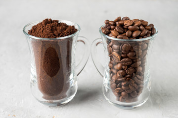 Two glass cups with coffee beans and ground coffee on light concrete background