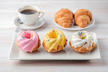 Three multi-colored cupcakes on a rectangular plate, cup of black coffee and plate with two croissants on a light table texture.