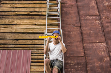 A man sits on the roof on a ladder with a building level