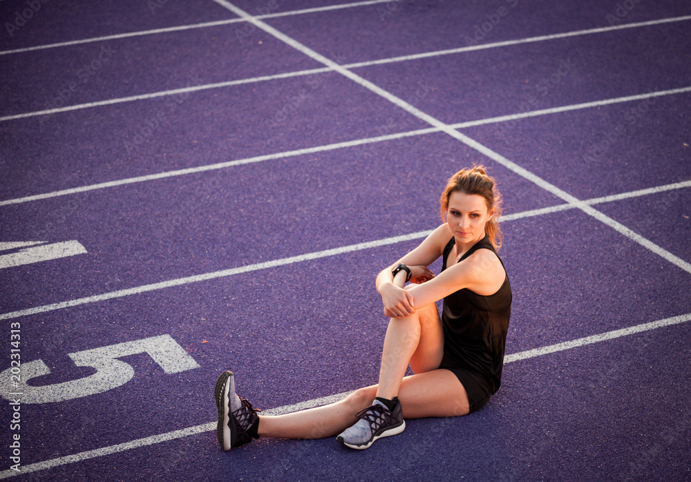 Poster Athletic woman sitting on running track and resting after training