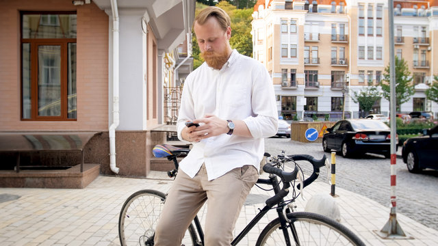 Photo of young man with beard leaning on old bicycle on street and typing message on smartphone