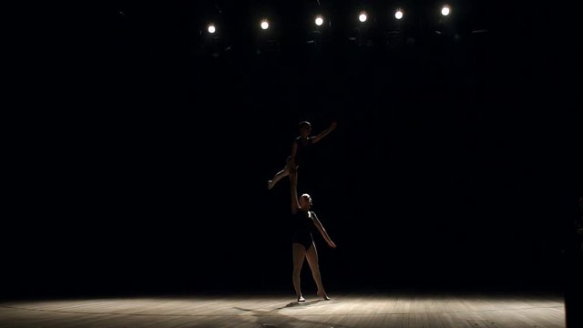 Two flexible girls gymnasts or ballerinas show a complex trick on stage in the light of white lights and smoke. The girl is standing on the shoulders of another girl. Circus trick. Slow motion.