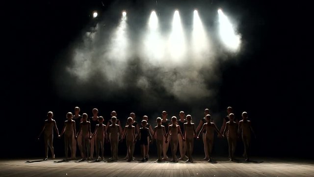 A very large group of children perform and dance on stage in the dark with smoke and concert lighting, they simultaneously bow at the end of the performance. Dancing. Ballet.