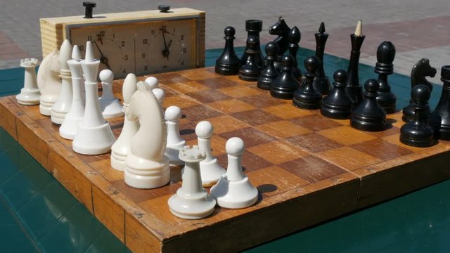 Black and white chess stand on the board, next to a vintage chess clock on street