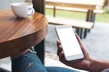 Mockup image of woman's hand holding white mobile phone with blank screen and coffee cup on wooden table in cafe