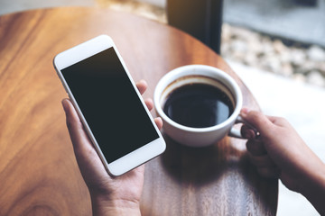 Mockup image of hands holding white mobile phone with blank black screen while drinking coffee in cafe