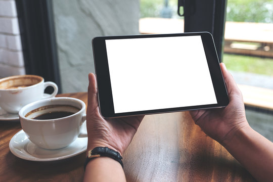 Mockup image of hands holding black tablet pc with white blank screen and coffee cups on table