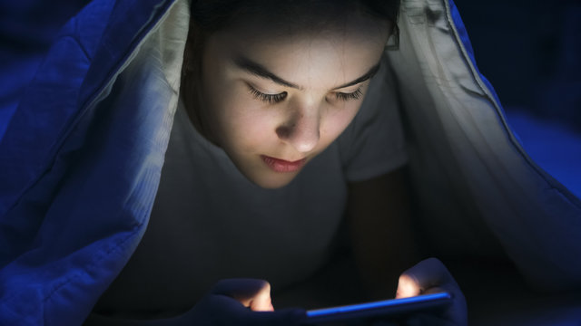 Closeup photo of girl in pajamas browsing internet on smartphone under blanket at night