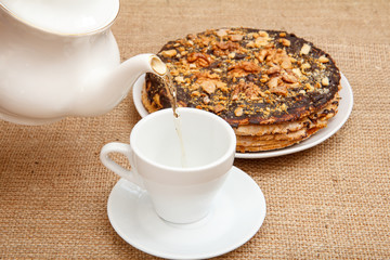 Tea is poured into a cup from porcelain teapot with homemade chocolate puff cake on the background