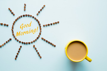 Yellow mug of coffee with milk on light pastel blue table from above. Sun created from brown beans. Inscription good morning. Empty place for inspirational, motivational text or quote. Top view.