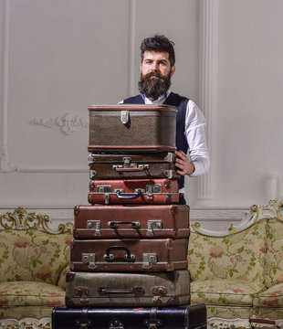 Macho elegant on strict face standing near pile of vintage suitcase. Luggage and travelling concept. Man, traveller with beard and mustache packing luggage before trip, luxury interior background.