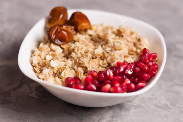 Cooked yummy warm oatmeal and heap of ripe fresh pomegranate seeds and three dried dates in white ceramic bowl on worn gray scratched concrete