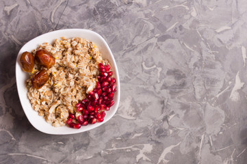 Cooked yummy warm oatmeal and heap of ripe fresh pomegranate seeds and three dried dates in white ceramic bowl on worn gray scratched concrete. Top view with copy space