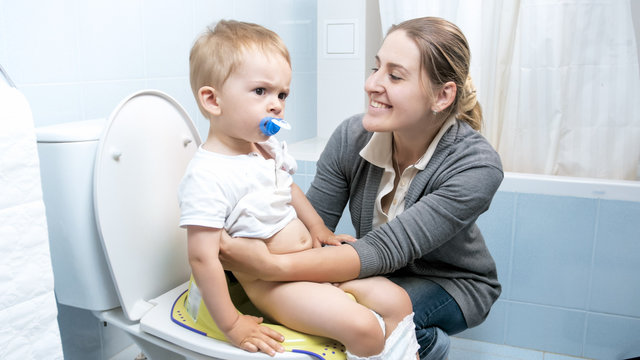 Portrait of young smiling mother seatting her toddler son on toilet