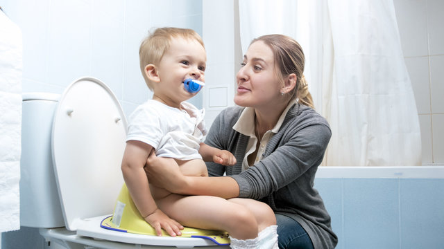 Cute smiling toddler boy sitting on toilet with young mother