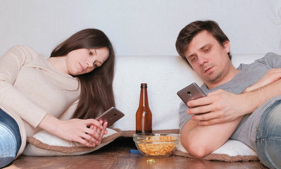 Couple man and woman browsing internet in their mobile phones drinking beer and eating chips.