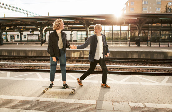 Cheerful young man pulling teenage girl on skateboard at railroad station platform