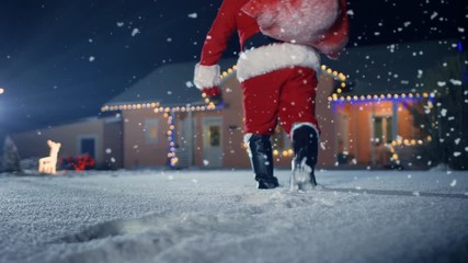 Santa Claus Carrying Red Bag over the Shoulder, Walks into Front Yard of the Idyllic House Decorated with Lights and Garlands. Santa Bringing Gifts and Presents. - Powered by Adobe