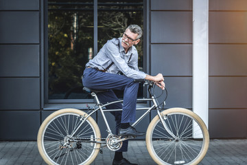 Modern lifestile - modern business. Side view of mature businessman looking over shoulder while riding on his bicycle.