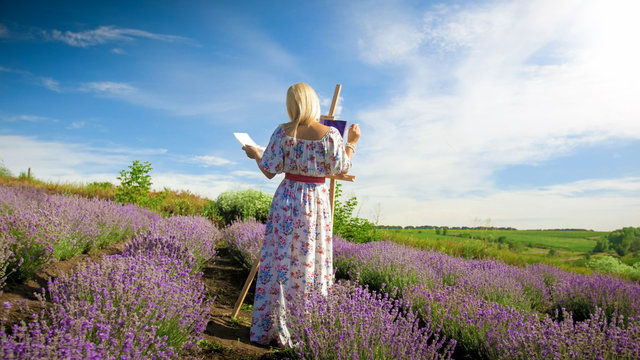 Rear view image of young woman in long dress painting picture of beautiful lavender field at Provence