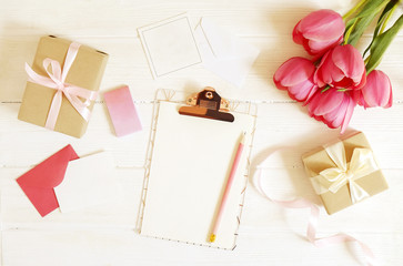 Feminine desktop composition with blank greeting card sheet clipboard, envelope, pink tulips bouquet, craft paper present wrap on white wood table background. Top view, flat lay, close up, copy space.