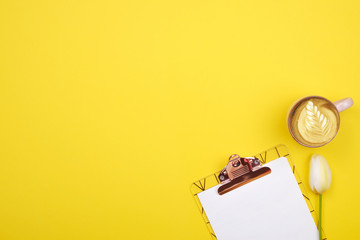 Feminine desktop composition with blank sheet clipboard, pencil, coffee cup, tulips bouquet on yellow background. Girlie workplace, flowers for mother's day. Top view, flat lay, close up, copy space.