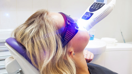 Portrait of young blonde woman sitting in dentist office while her teeth being whitened with UV light