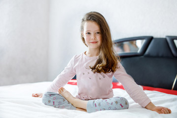 Obraz na płótnie Canvas Adorable happy little kid girl after sleeping in his white bed in colorful nightwear. School child making baby yoga exercises