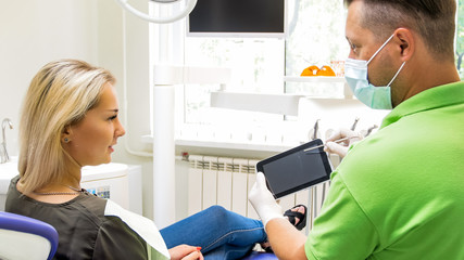 Portrait of male orthodontist talking to his patient about teeth treatment and showing digital tablet. Place for your text or image on screen