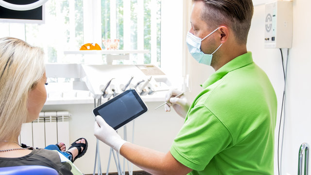 Portrait of male dentist holding digital tablet and showing it to his patient. Place for your text or image on screen