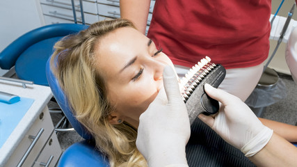 Closeup image of dentist in latex gloves holding teeth color tone samples at patients teeth