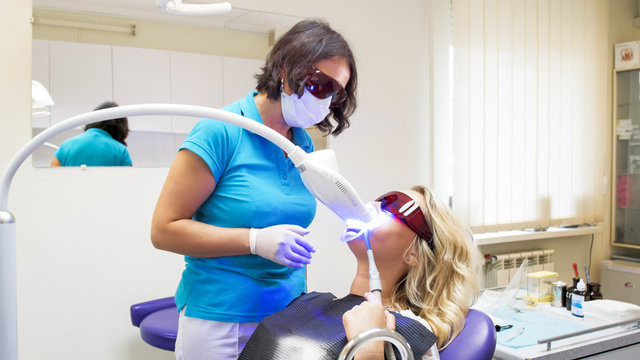 Female dentist in protective eyeglasses looking at patient during teeth whitening procedure with UV light