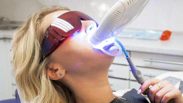 Closeup image of young woman in protective glasses sitting in dentist chair during teeth whitening procedure