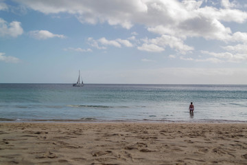 Fototapeta na wymiar Woman bathing in the sea, yellow sand beach, with sailboat on the horizon, blue sky with clouds. Fuerteventura, Canary Islands.