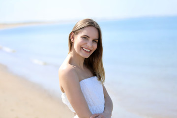 Fototapeta na wymiar Hübsche blonde Frau in einem weissen Kleid steht lachend am Strand und Meer in Hintergrund