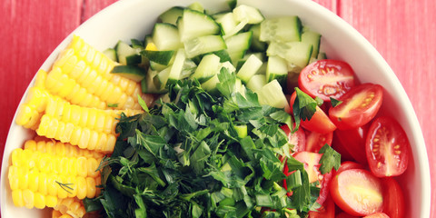 White plate with cut vegetables for a vegan salad on the red wooden table. Colorful photo. Corn, cherry tomatoes, bell pepper, onion, cucumber and garden herbs. Wide photo. Top view.