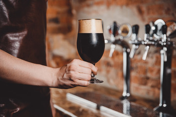 Barman holds in his hand dark unfiltered cold beer is poured into glass beaker against background of taps for bottling.