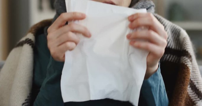 Close up of the young unwell caucasian woman in the warm scarf and a plaid sneezing in the napkin as she having a cold. At home. Inside