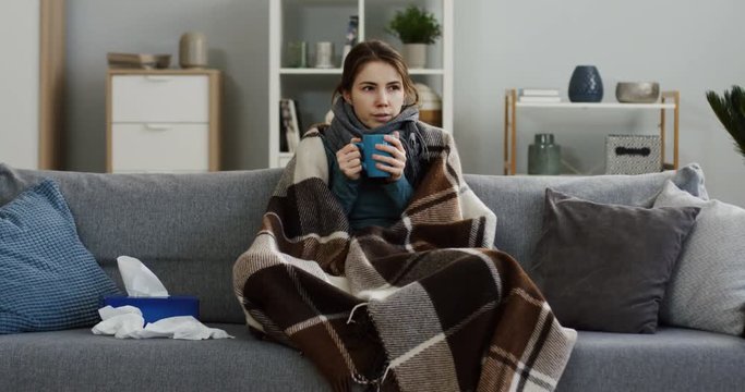 Unwell caucasian young woman sitting on the gray sofa in the warm plaid and drinking hot tea as she having an influenza. At home. Indoors