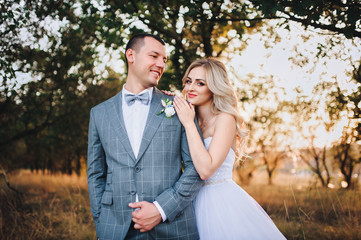 Fototapeta premium Lovely and stylish newlyweds are hugging and smiling at the background of the chic nature at sunset. The bride with curly hair leaned against the shoulder of her beloved bridegroom. 
